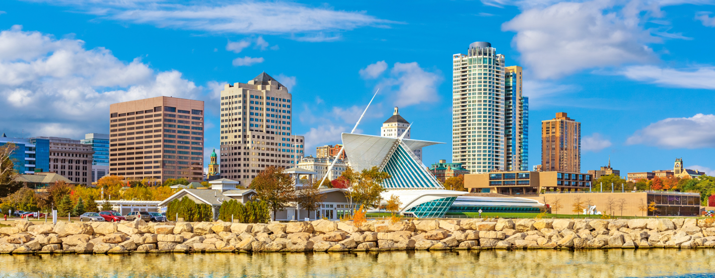 Milwaukee downtown skyline set against blue sky is reflected in the waters of Lake Michigan in foreground.