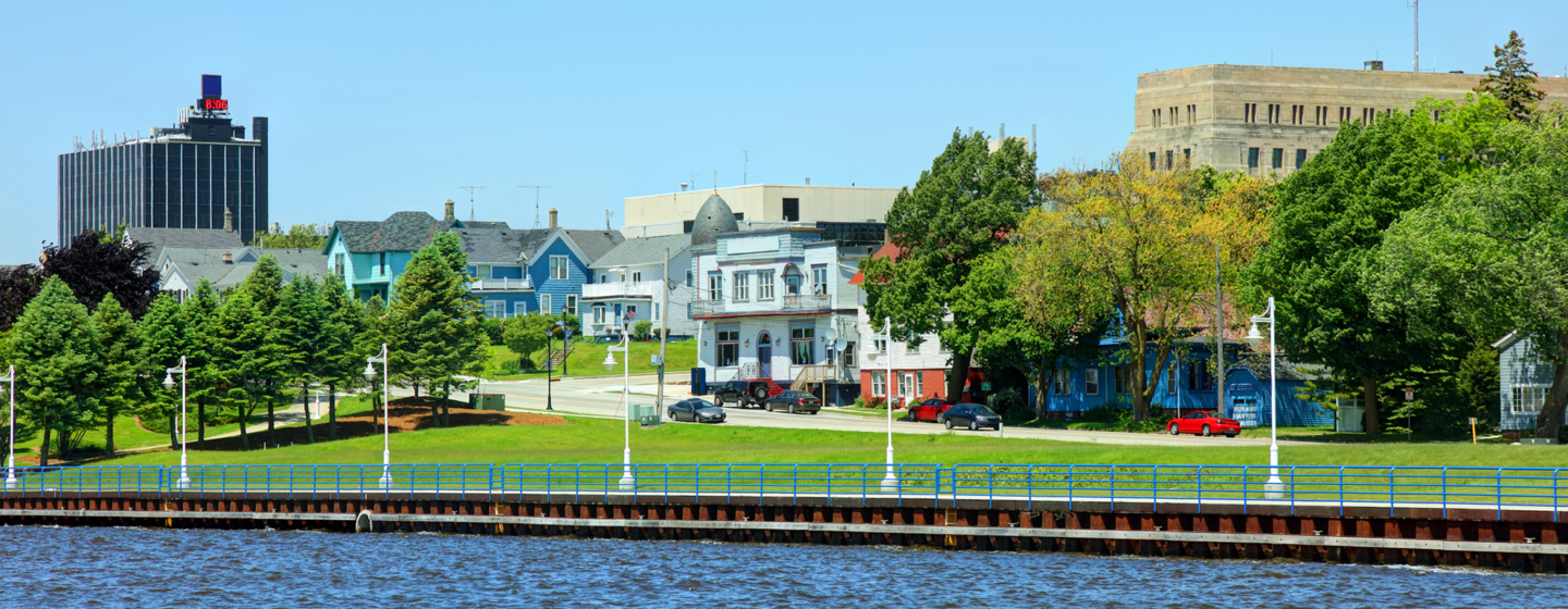 Trees and buildings line Sheboygan's downtown area on sunny summer day with body of water in foreground.