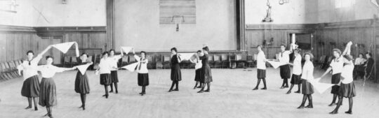 Group of students at gymnasium exercising in semicircle with white scarves.