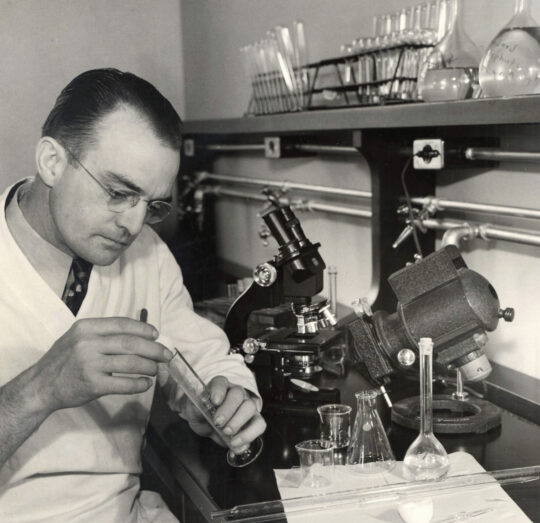 Paul Phillips working with long granulated cylinder at table covered with microscopes and other laboratory glassware.