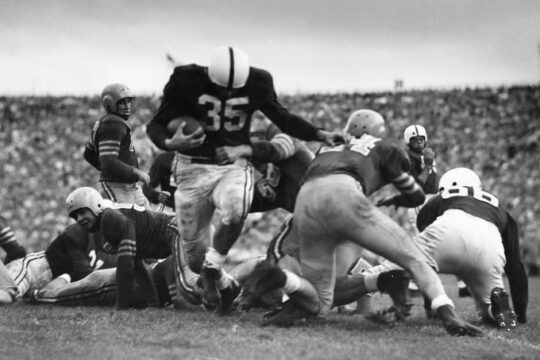 Alan Ameche carrying football attempting to break tackle while running through line of players during football game.