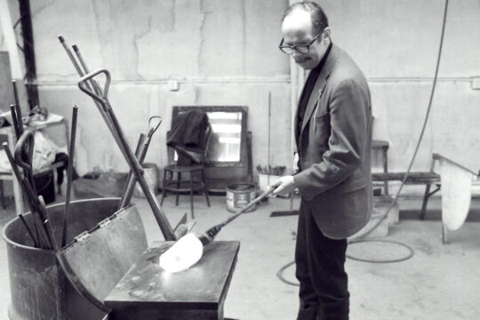 Harvey Littleton in studio rolling piece of hot glass using long pipe on bench.