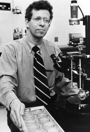 Howard Temin in laboratory standing beside microscope and tray of transparent lidded dishes.