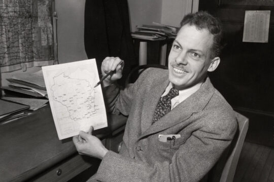 Frederic Cassidy at desk holding up and pointing to Wisconsin state map on sheet of paper.