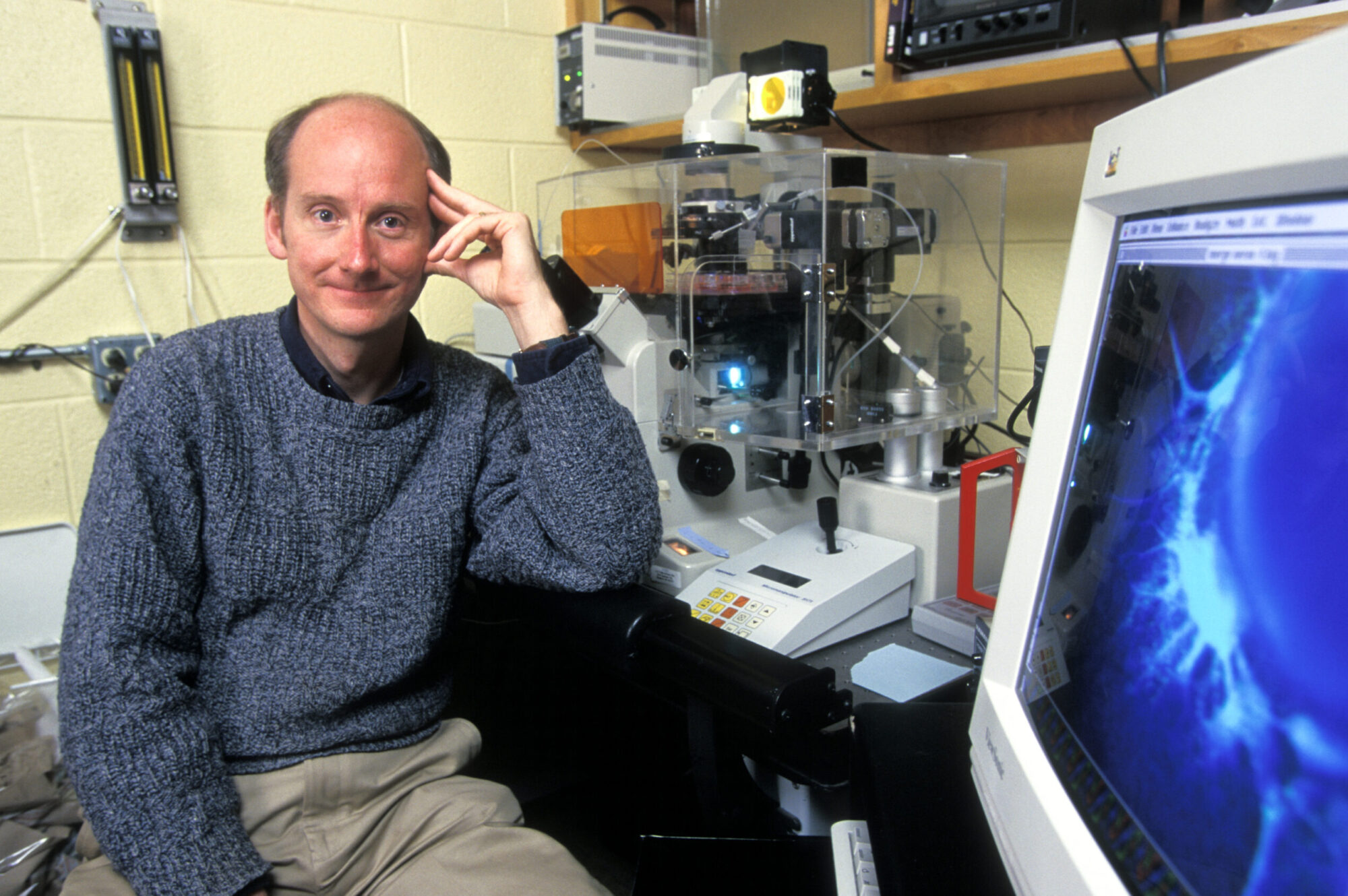 James Thomson posing beside lab equipment and computer displaying image of blue stem cell neurons in research laboratory.