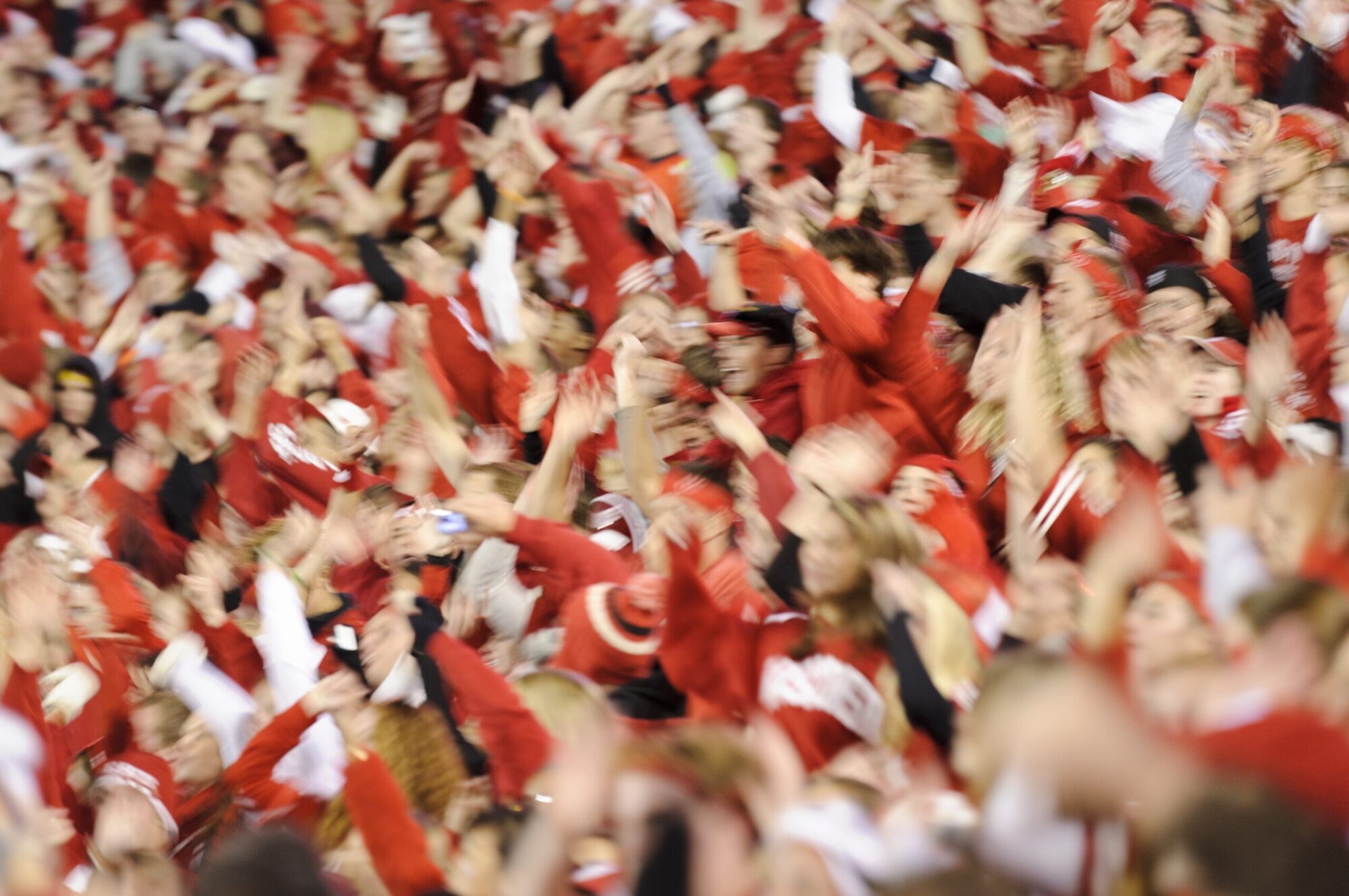 Badger football fans dressed in red cheering and jumping around with hands in air at Camp Randall Stadium.
