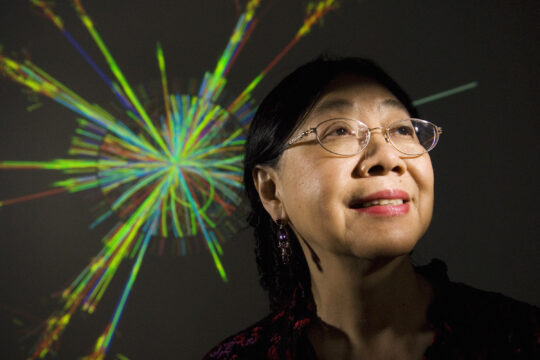 Sau Lan Wu posing against projection of colorful computer-generated simulation depicting paths of subatomic particles produced by colliding beams of protons.