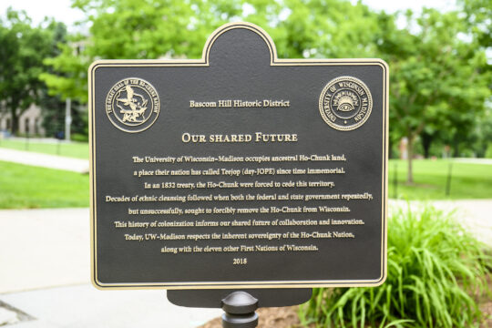 Black and gold Our Shared Future plaque positioned at top of Bascom Hill displaying trees, greenery, and sidewalks in background.