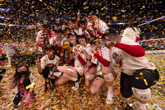 Gold confetti flies around celebratory members of UW women's volleyball team as they pose with the 2021 NCAA Division I Volleyball Championship trophy.