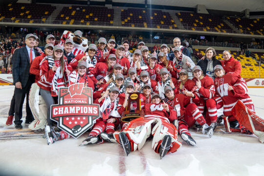 UW Badgers women's hockey team and coaching staff celebrating their 2023 National Championship victory on the ice.