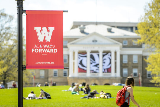 Red banner on Bascom Hill featuring "All Ways Forward" slogan with students enjoying sunny day.