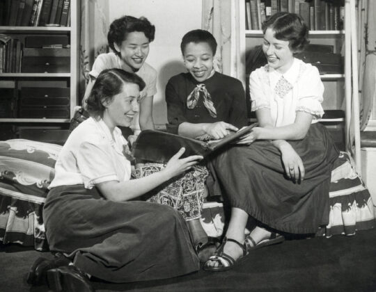 Four smiling students sitting together in room at Groves Housing Cooperative looking at book.