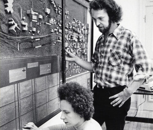 Person crouched down using fingers to explore three-dimensional campus map attached to wall while another person observes.