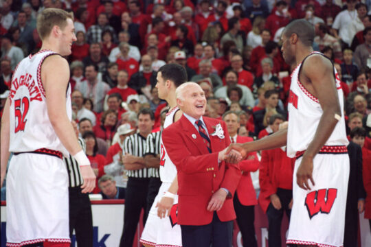 Senator Herb Kohl standing on court at Kohl center shaking hands with basketball player as crowd of fans watch.