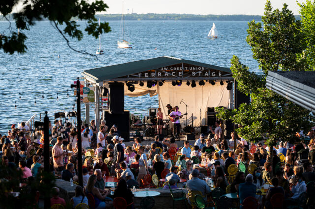 Large group of people listening to live music at Memorial Union Terrace stage with Lake Mendota and sailboats in background.