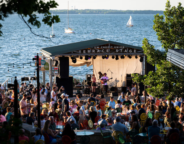 Large group of people listening to live music at Memorial Union Terrace stage with Lake Mendota and sailboats in background.