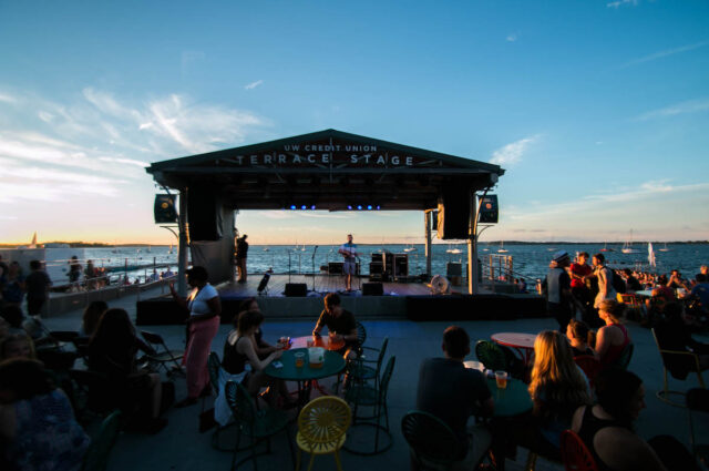 Crowd of people gathered at Memorial Union Terrace overlooking Lake Mendota and watching live music performance of singer playing ukulele.