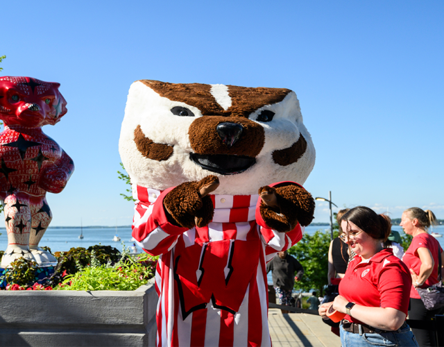 Bucky Badger outdoors at terrace with Bucky statue positioned in background.