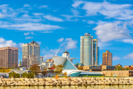 Milwaukee downtown skyline set against blue sky is reflected in the waters of Lake Michigan in foreground.