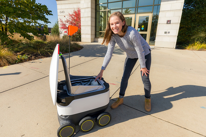 A photo of the white starship delivery robot in front of a residence hall, with a student leaning down to touch the top.