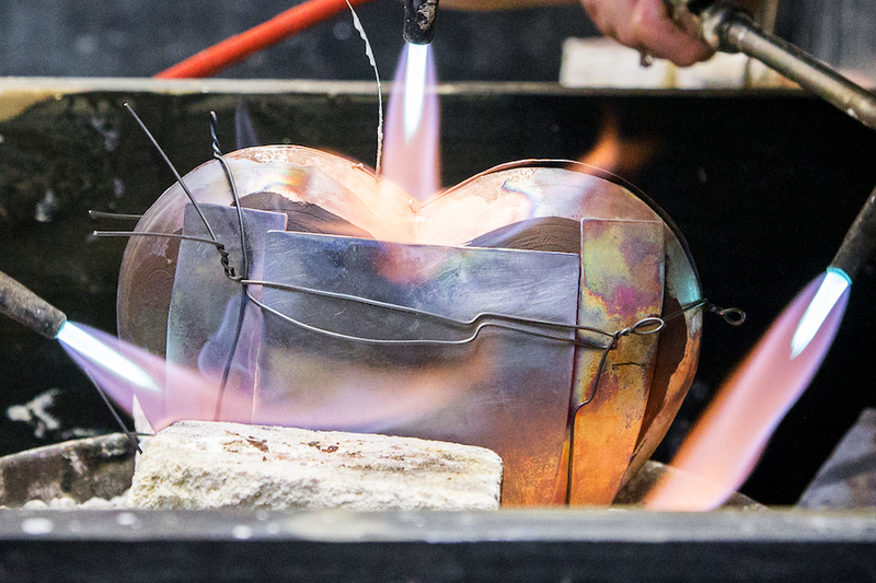 A photo of a metal heart being welded together using 3 torches.