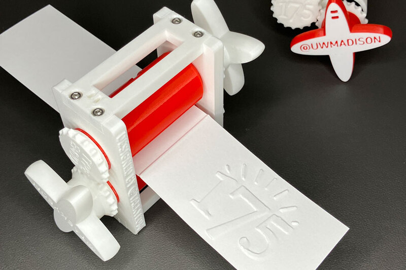 A photo of a 3D printed embosser with the 175th Anniversary Logo, with a paper running through it.