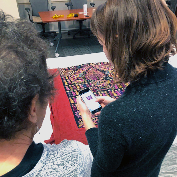photo of two people taken from behind looking over their shoulders and they are looking at a textile on a table and taking a photo of it with their phone.