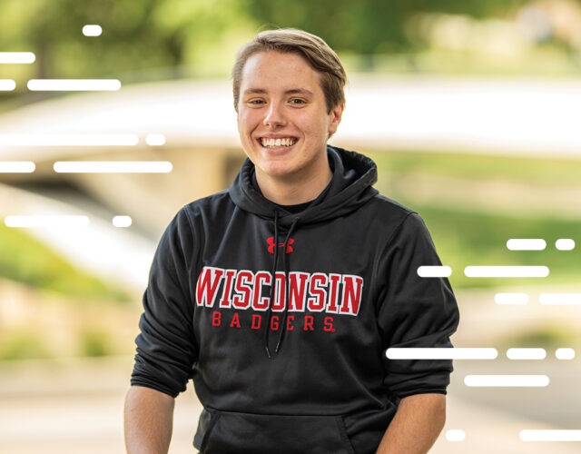 Student wearing Wisconsin Badgers black hoodie smiling at camera against blurred background.