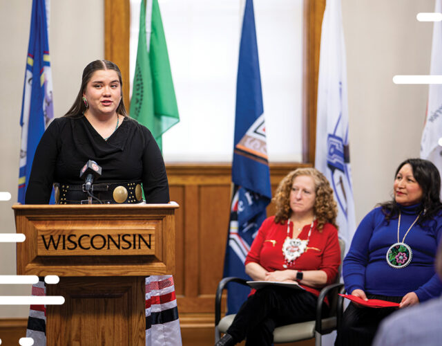 UW student with the Odaawaa-zaaga’iganing tribe speaking at podium with Chancellor Mnookin in background.