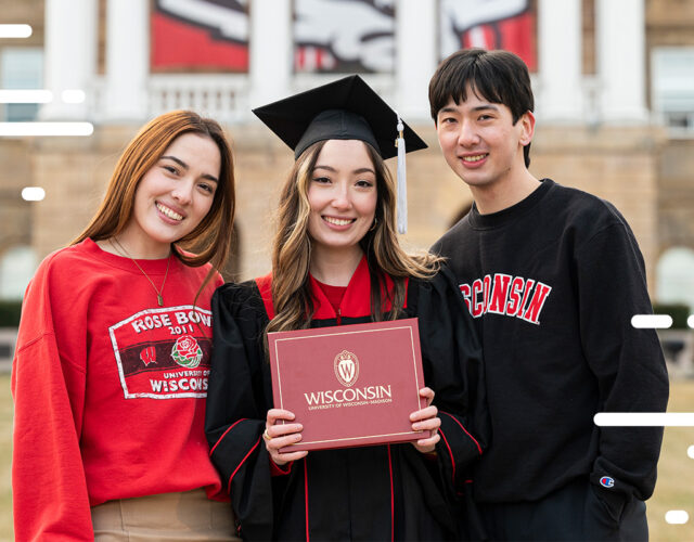 UW–Madison graduate wearing cap and gown holding graduation diploma standing next to siblings in front of Bascom Hall.