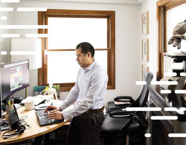 Person standing at desk in office typing on keyboard looking at computer screen.