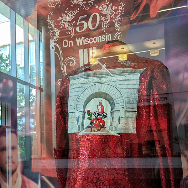 A red sequined jacket with a Camp Randall and Bucky in a marching band uniform holding a rose patch on the back. Behind the jacket is a cropped photo of Mike Leckrone directing at the spring show. In the foreground glass reflections are seen.