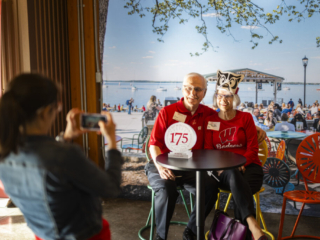 175 Celebration in Appleton Two alums in red University of Wisconsin–Madison attire posing for photo in front of backdrop sitting on sunburst terrace chairs.