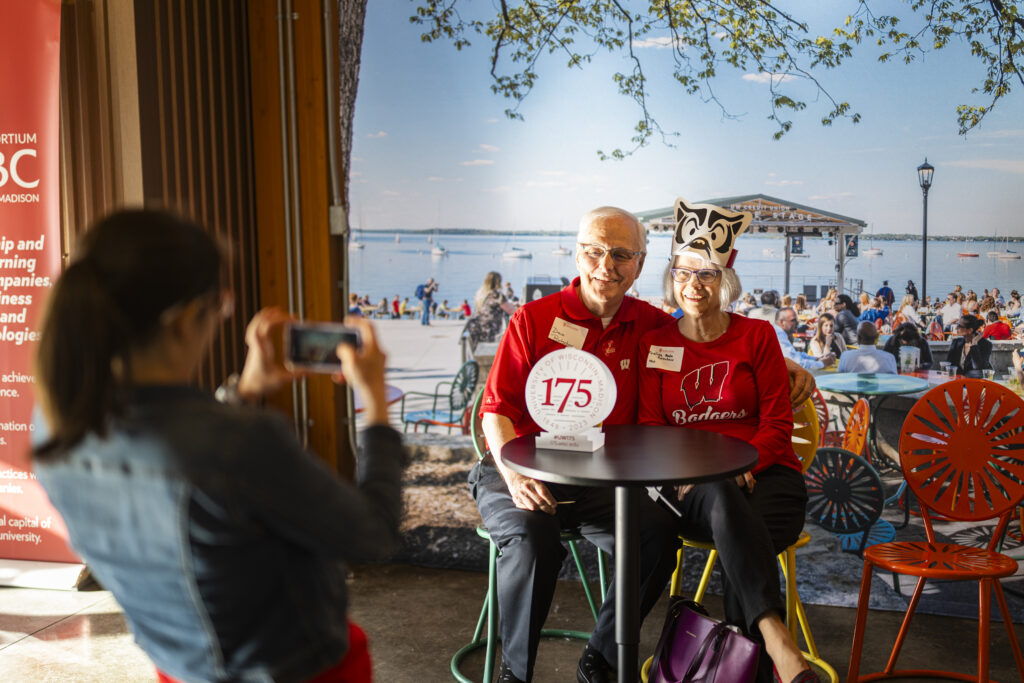 Two alums in red University of Wisconsin–Madison attire posing for photo in front of backdrop sitting on sunburst terrace chairs.
