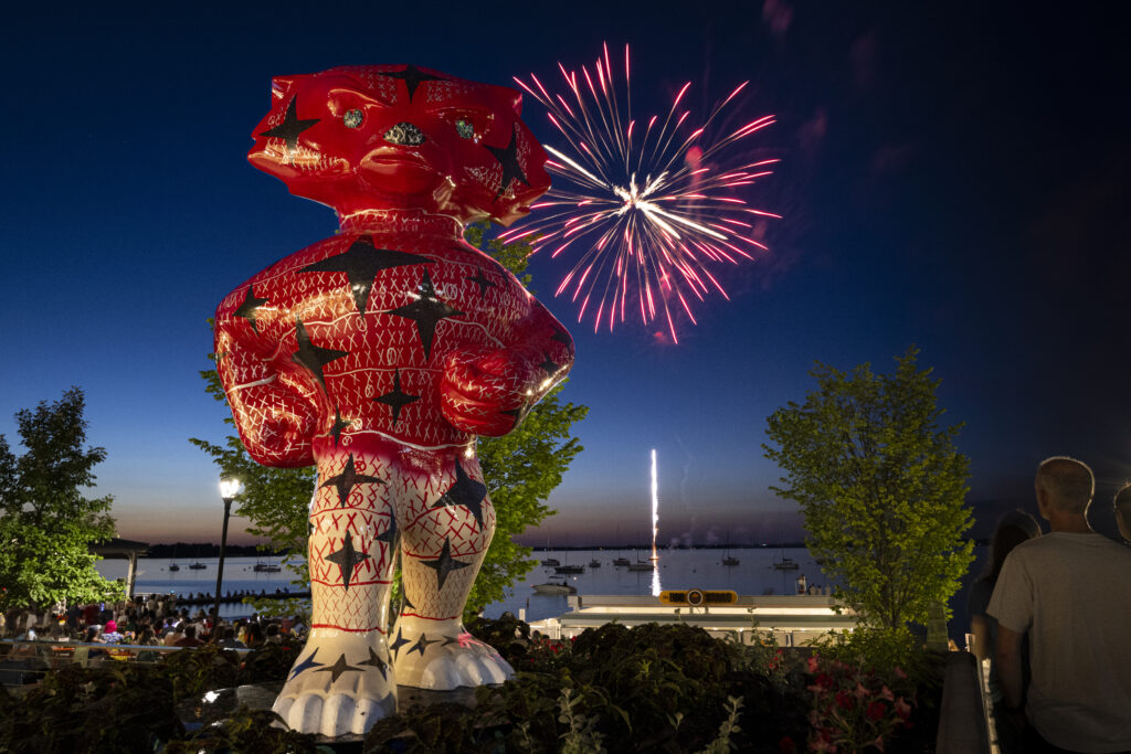 Bucky Badger statue in foreground with fireworks booming over lake during summer evening in background.