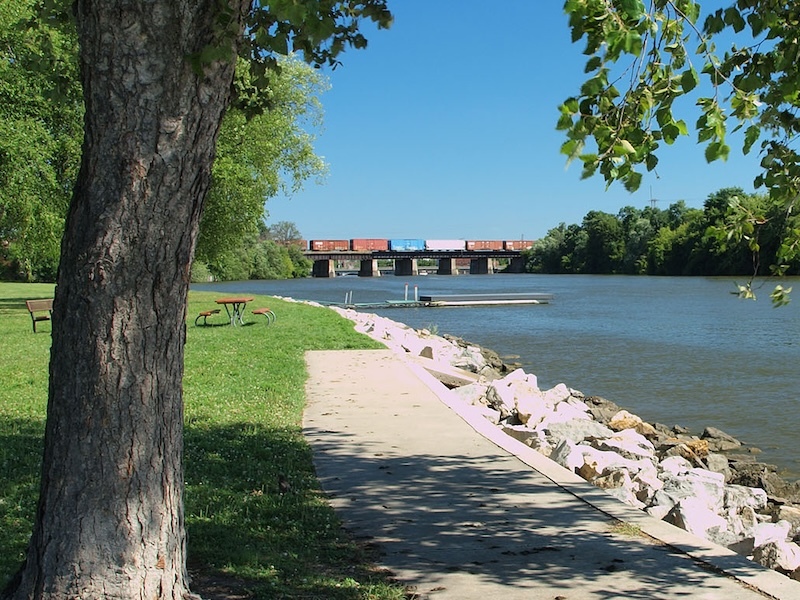 Riverfront lined by rocks, concrete path, picnic table and dock in foreground and trees in background.