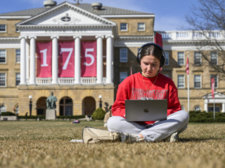 175 Banners - Bascom Hill Undergraduate student sitting on ground cross-legged working on laptop wearing headphones in front of Bascom Hall decorated with a 175 banner.