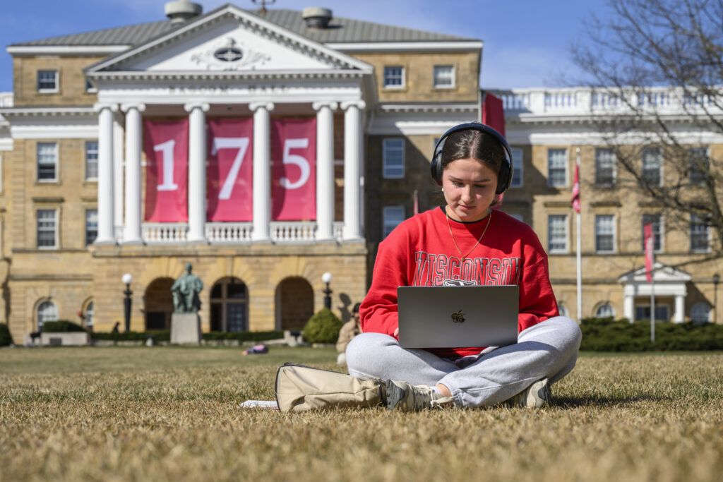 Undergraduate student sitting on ground cross-legged working on laptop wearing headphones in front of Bascom Hall decorated with a 175 banner.
