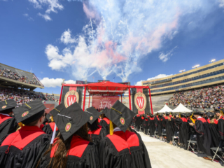 2024 Spring Commencement Graduates standing shoulder-to-shoulder in red and black cap and gowns facing stage with fireworks set off in background.
