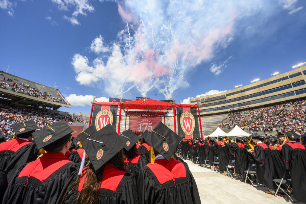 Graduates standing shoulder-to-shoulder in red and black cap and gowns facing stage with fireworks set off in background.