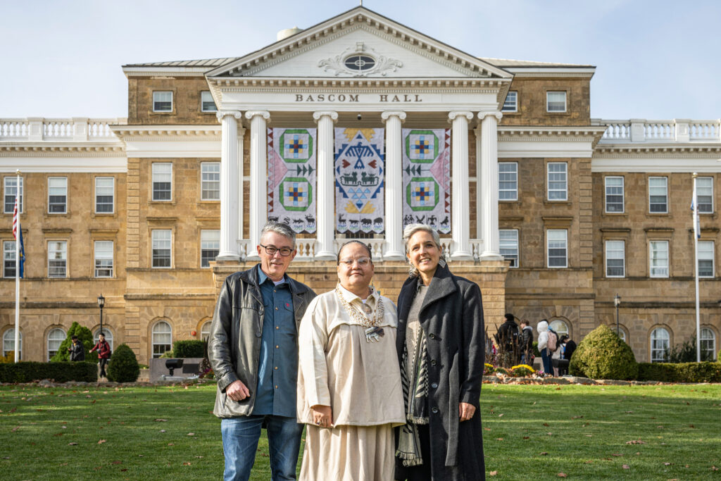 Three people posing for photo in front of Ho-Chunk banners hanging between building columns of Bascom Hall on autumn morning.
