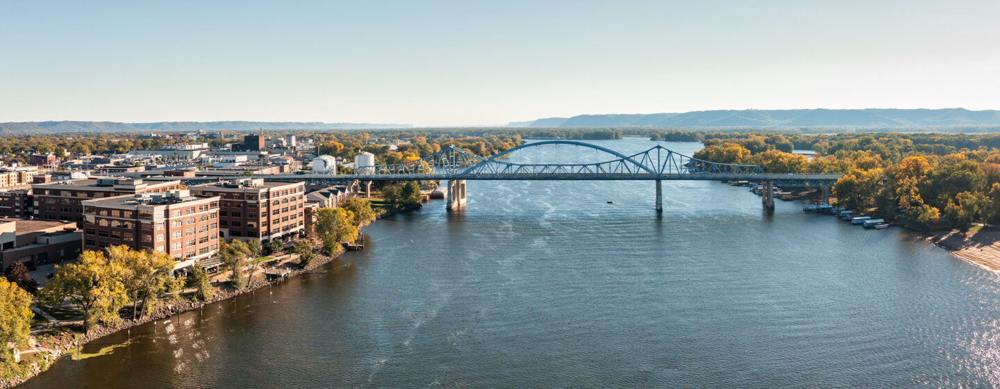 Arched bridge passing over river with city buildings on one side and trees on other side.