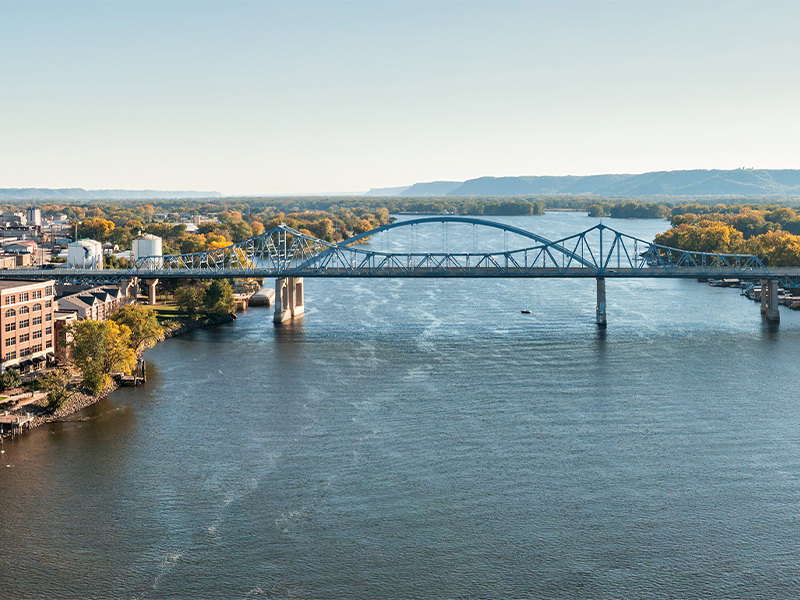 Arched bridge passing over river with city buildings on one side and trees on other side.