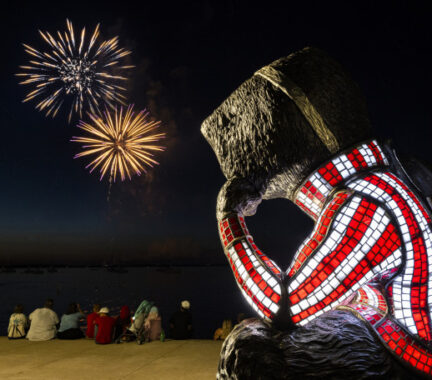 A statue of Bucky Badger is posed sitting with his cheek resting on his hand. He is looking away toward Lake Mendota where a fireworks display lights the sky.