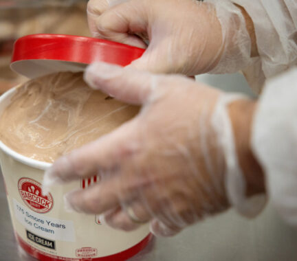 In a close-up phono, a person places a red lid on a container of ice cream labeled 175 S'more Years. The person is wearing clear food safety gloves.