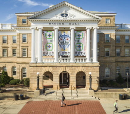 A view of the front facade of Bascom Hall taken from a drone. On a sunny day, a few people walk across the brick and concrete path in front of the building. The four panels of the Seed by Seed banner hangs between tall, white columns above the building's main entrance. The banner has been printed with a texture resembling beadwork and contains symbols and colors representing traditions of the Ho-Chunk Nation. Four green rings represent the four lakes of Teejop, the name the Ho-Chunk give the land now occupied by UW–Madison. Inside each ring, square patches in light blue, dark blue yellow and red represent the reflections of light on the water at different times of day. Two large pink triangles on either side of the banner represent flowers, with green stems and triangular leaves leading to the center panel. On the center panel, a large diamond made of small blue triangles frames a blue thunderbird, which is flanked by two red, abstract W's, representing UW–Madison. Below the thunderbird are two green water spirits, which resemble four-legged animals with very long tails. Below the water spirits are six light blue triangles representing water. Above and below the large diamond frame are bursts of yellow beading, representing the sun. Along the bottom border of the banners are stylized animal symbols of the twelve clans of the Ho-Chunk Nation, and beneath each animal is a traditional Ho-Chunk flower motif in blue and green.